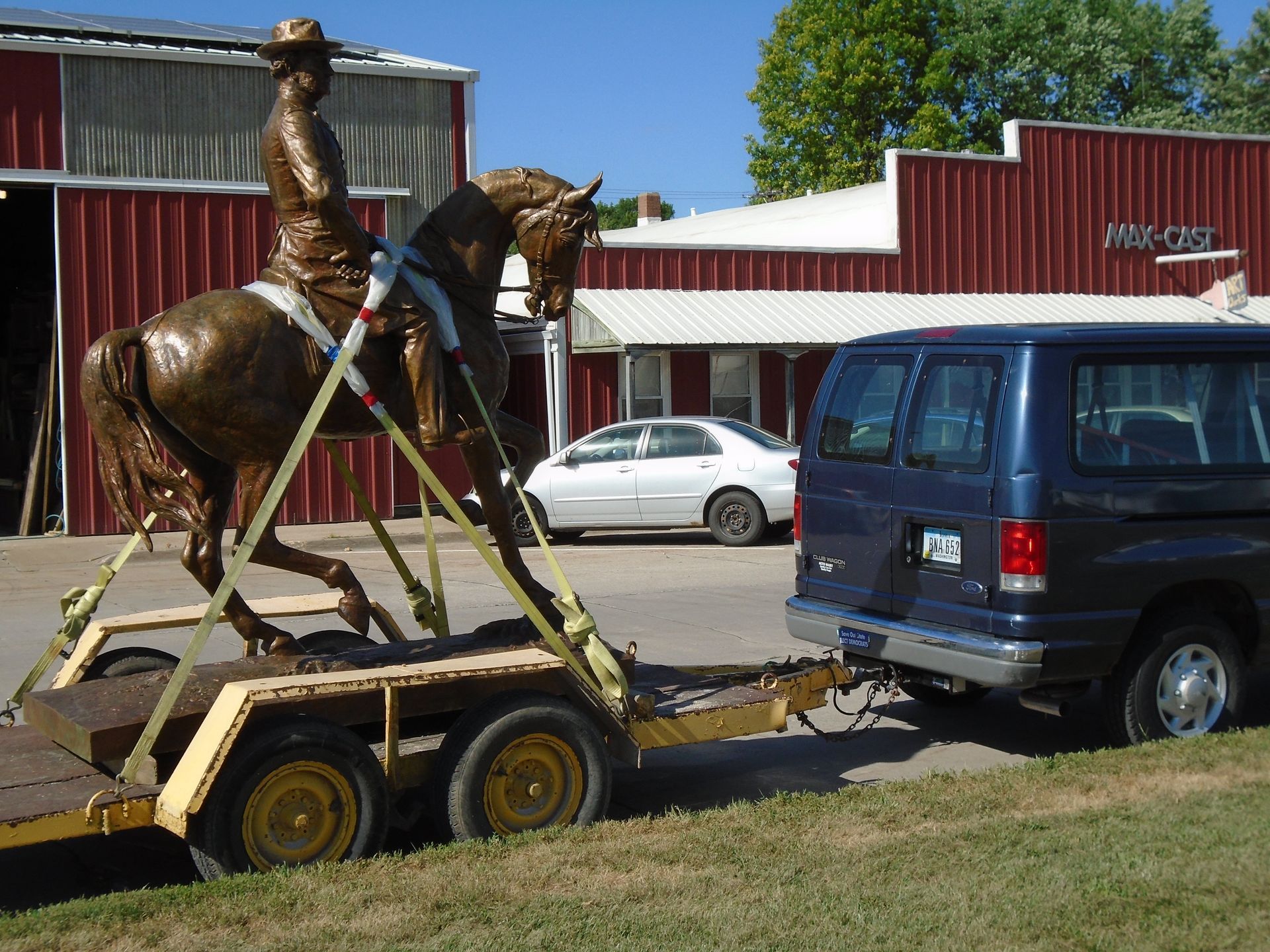 Bronze cowboy statue on a trailer being towed beside a blue van in front of a red barn