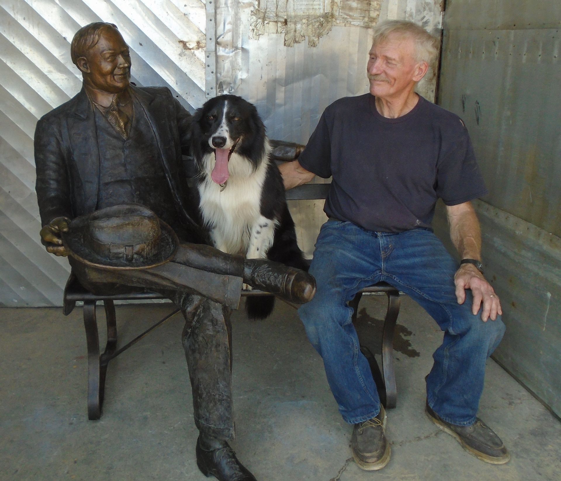 Man sitting beside a bronze statue on a bench, with a black-and-white dog between them, indoors.