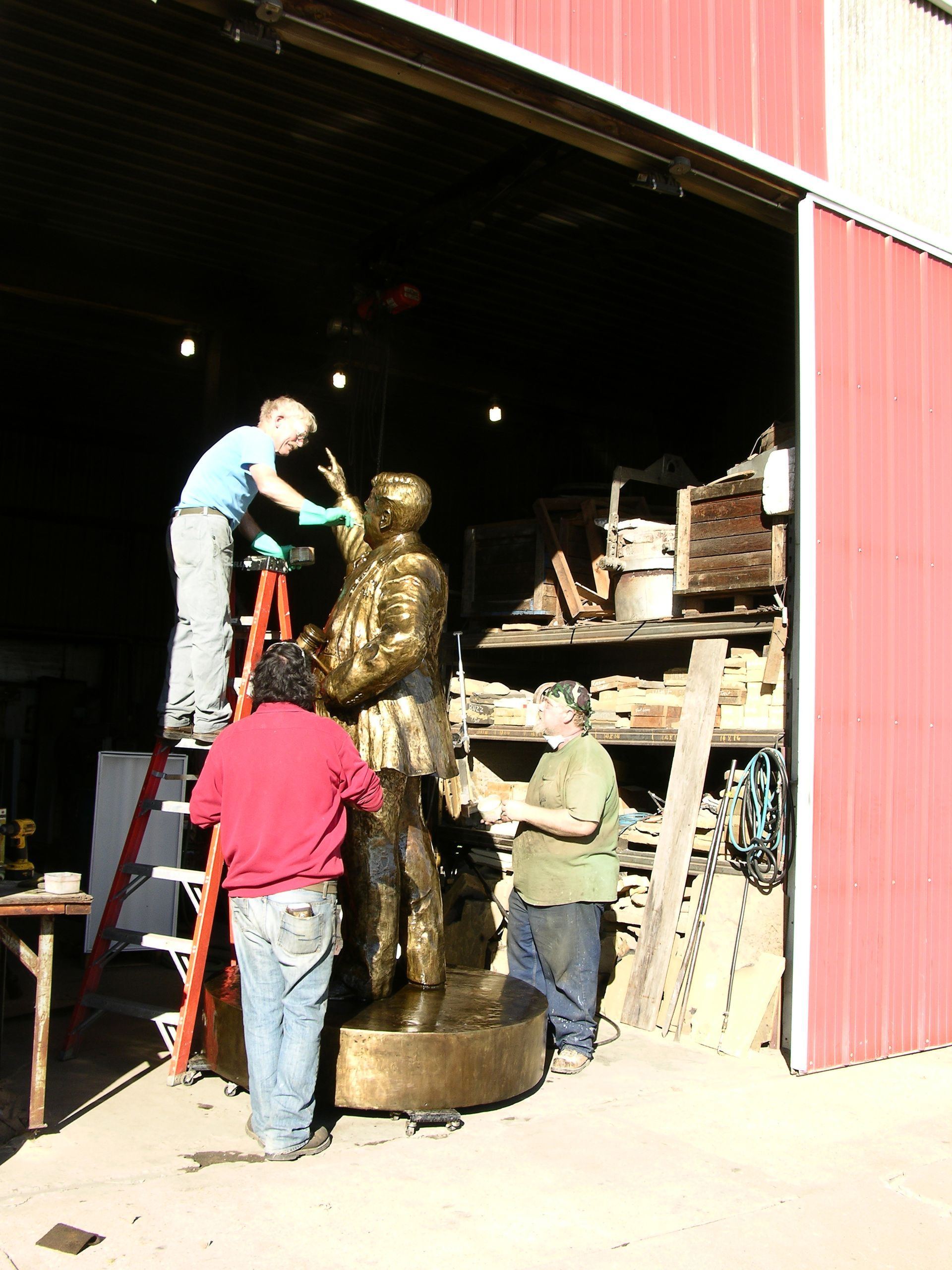 Workers restoring a gold statue in a warehouse, with one on a ladder and others nearby.