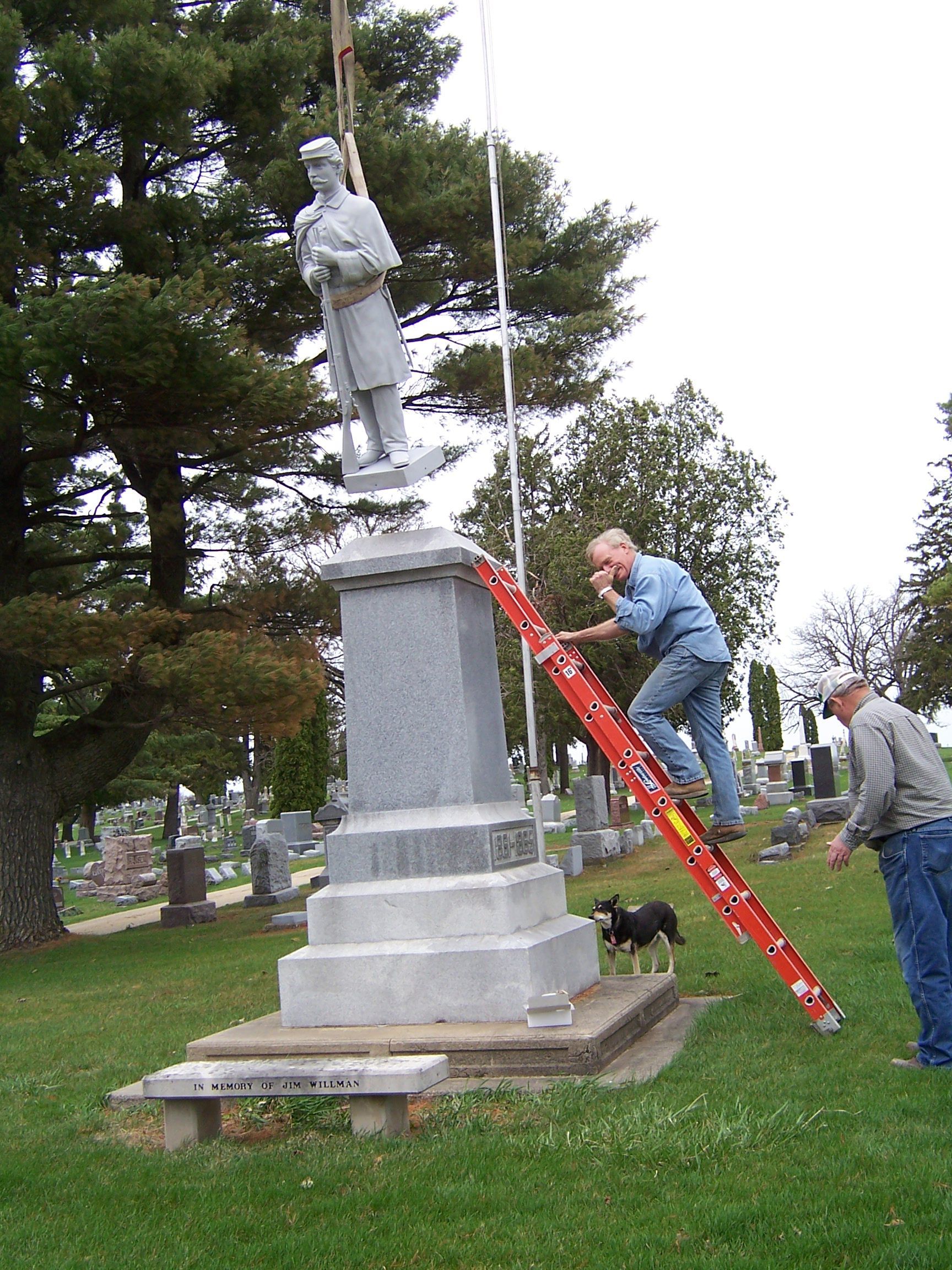 Two men using a red ladder to work on a statue atop a stone monument in a grassy cemetery