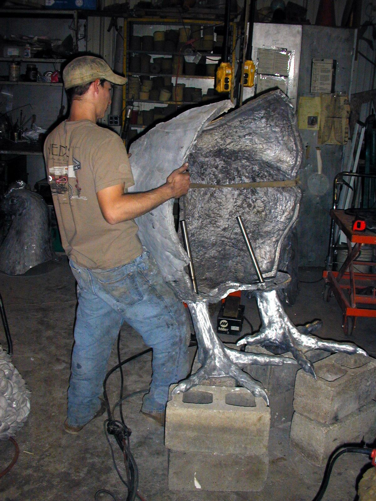 Person shaping a large silver sculpture in a workshop, standing beside cinder blocks and tools.