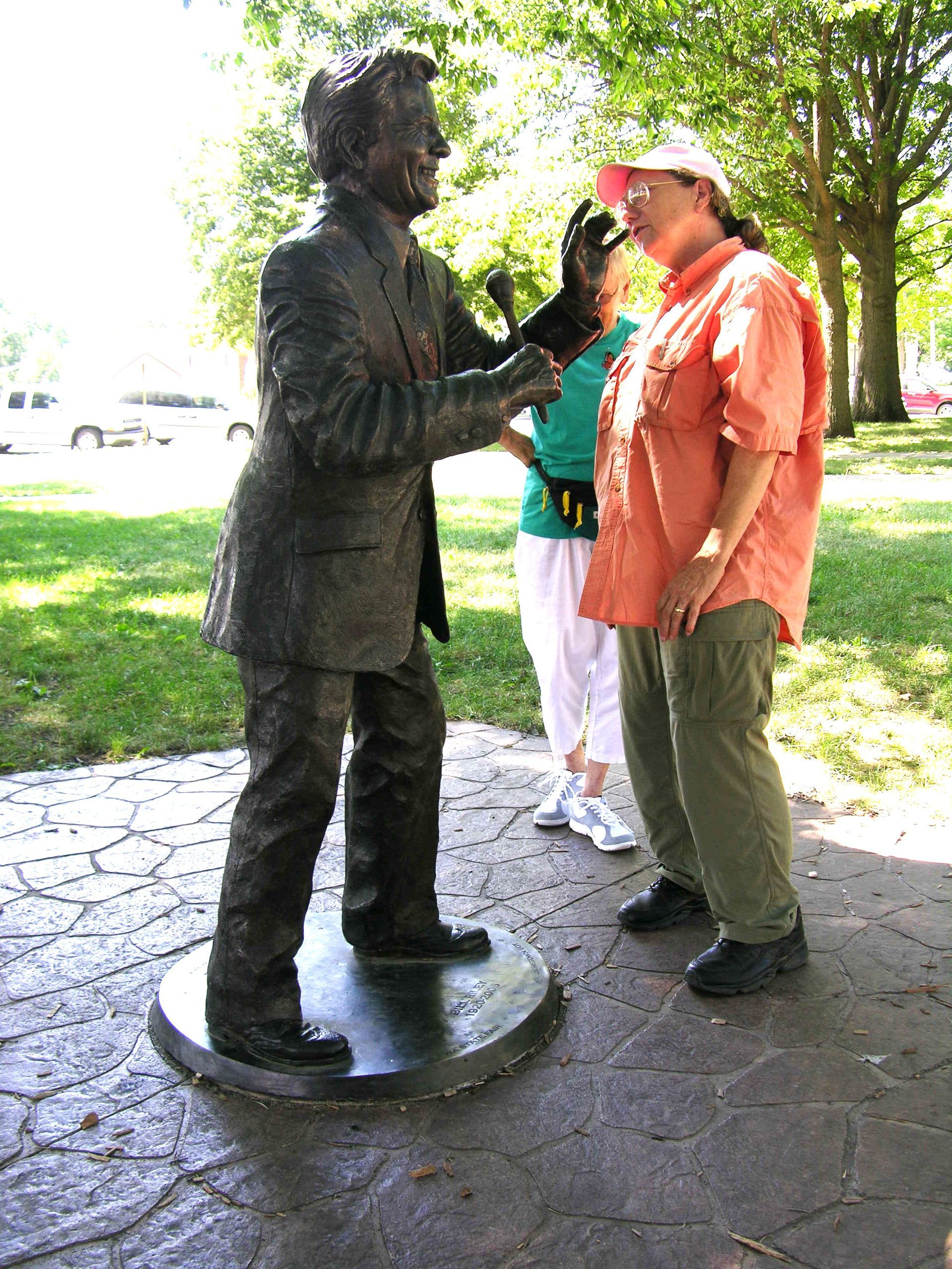 Bronze statue reaching toward a man in orange shirt and cap in a park walkway