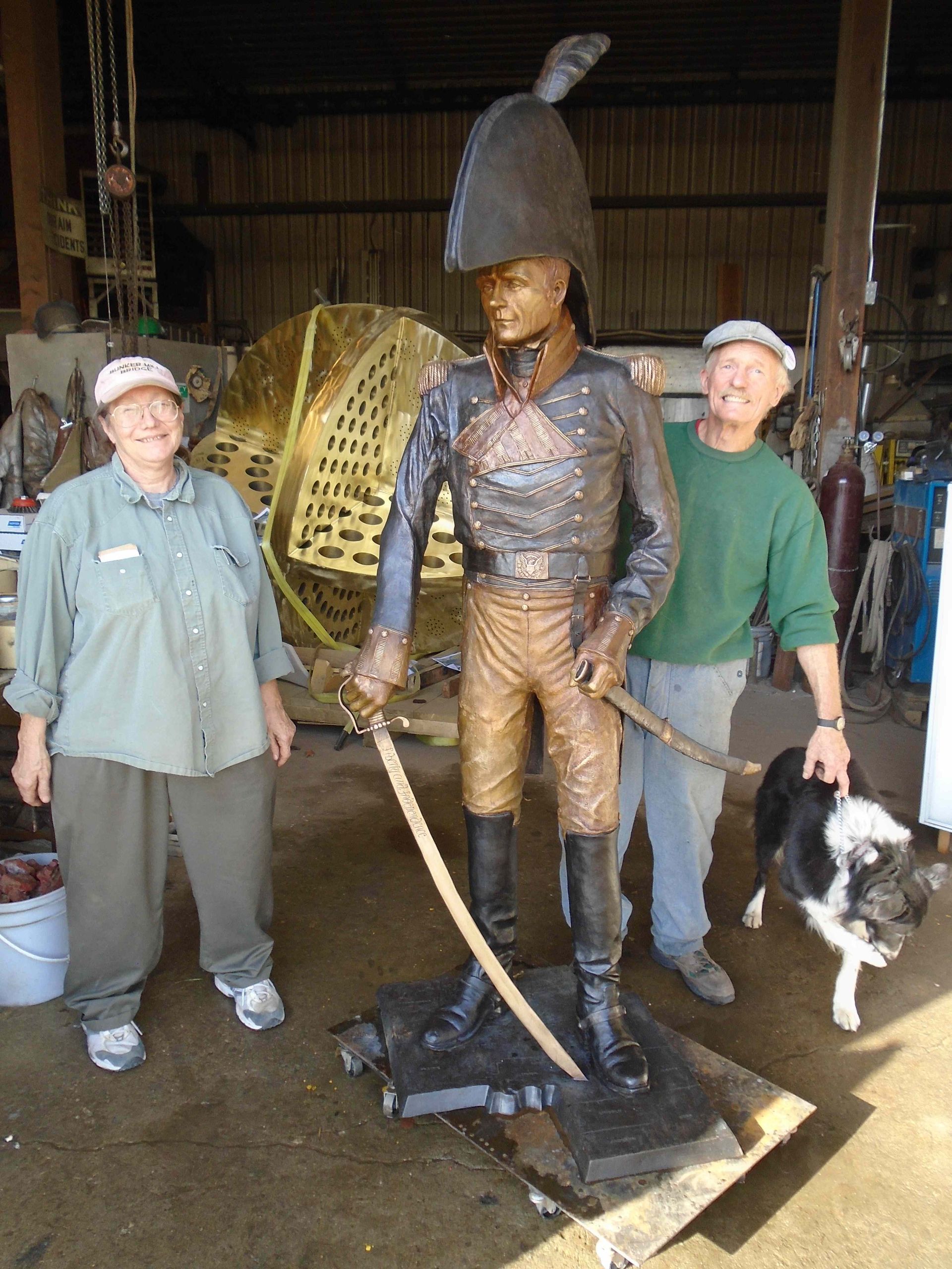 Two people and a dog stand beside a bronze statue in a workshop-like setting.