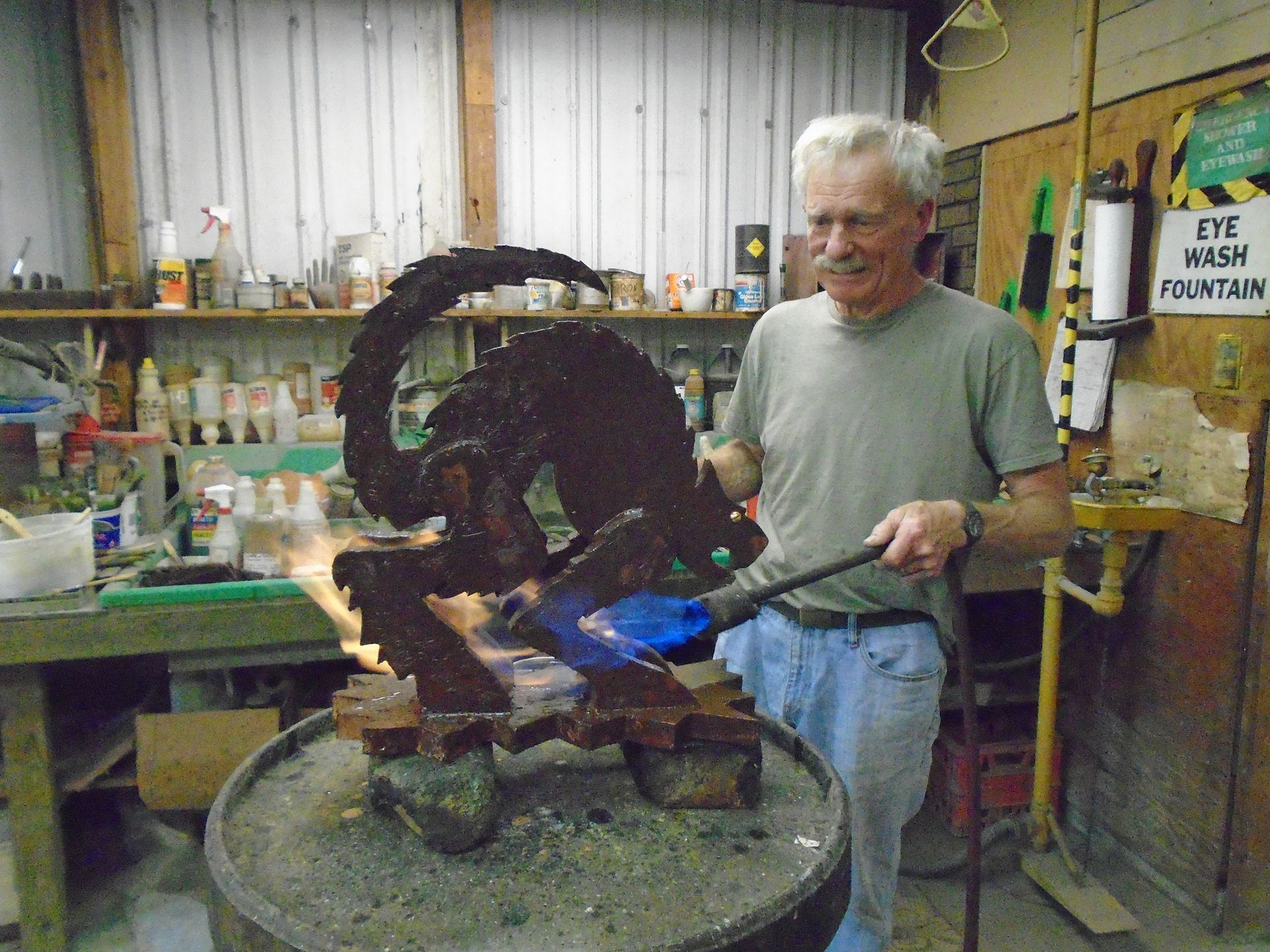 Man shaping a dark metal sculpture with a blue torch in a cluttered workshop