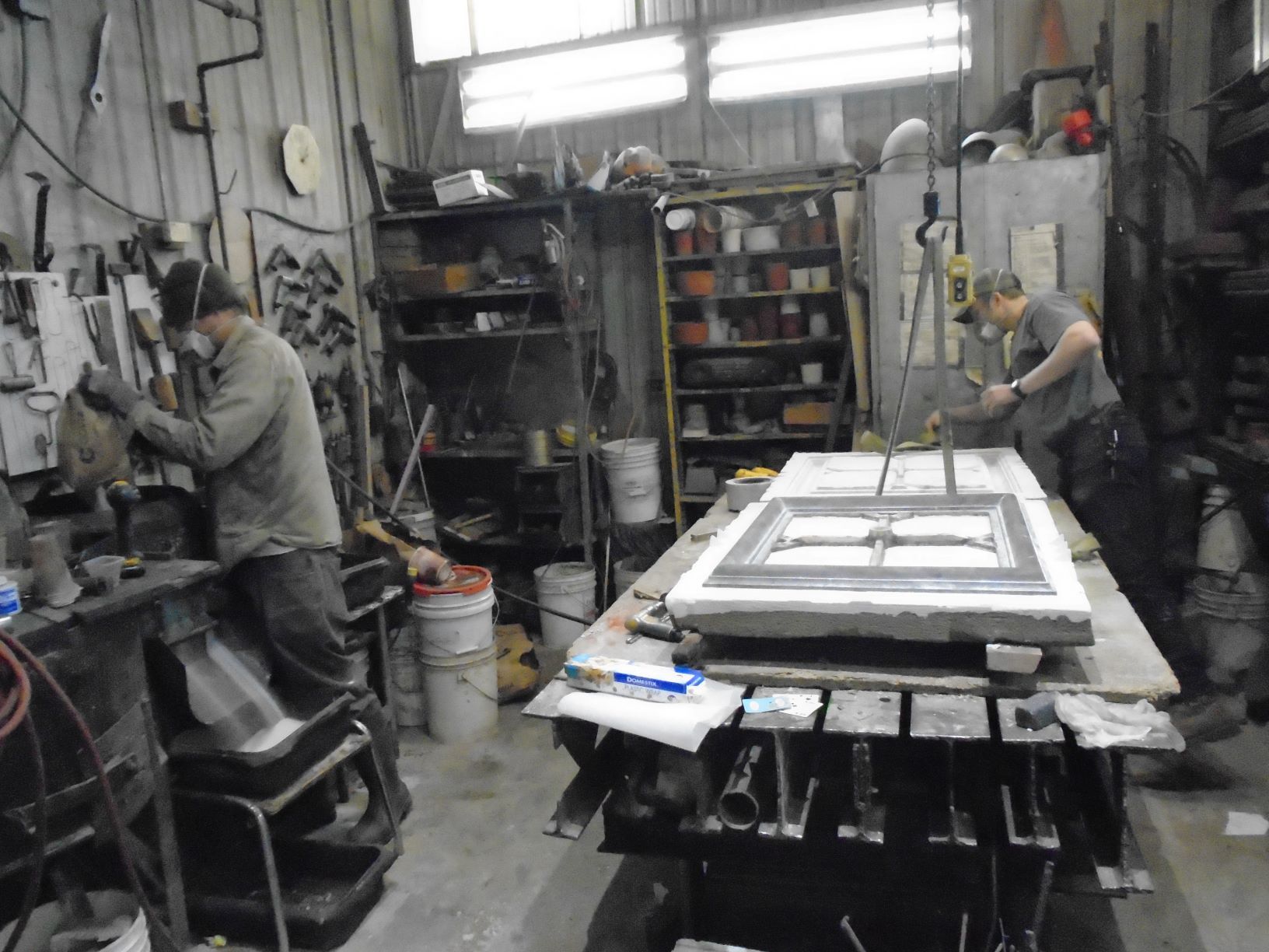 Two workers in a cluttered workshop, handling a large framed panel on a table.