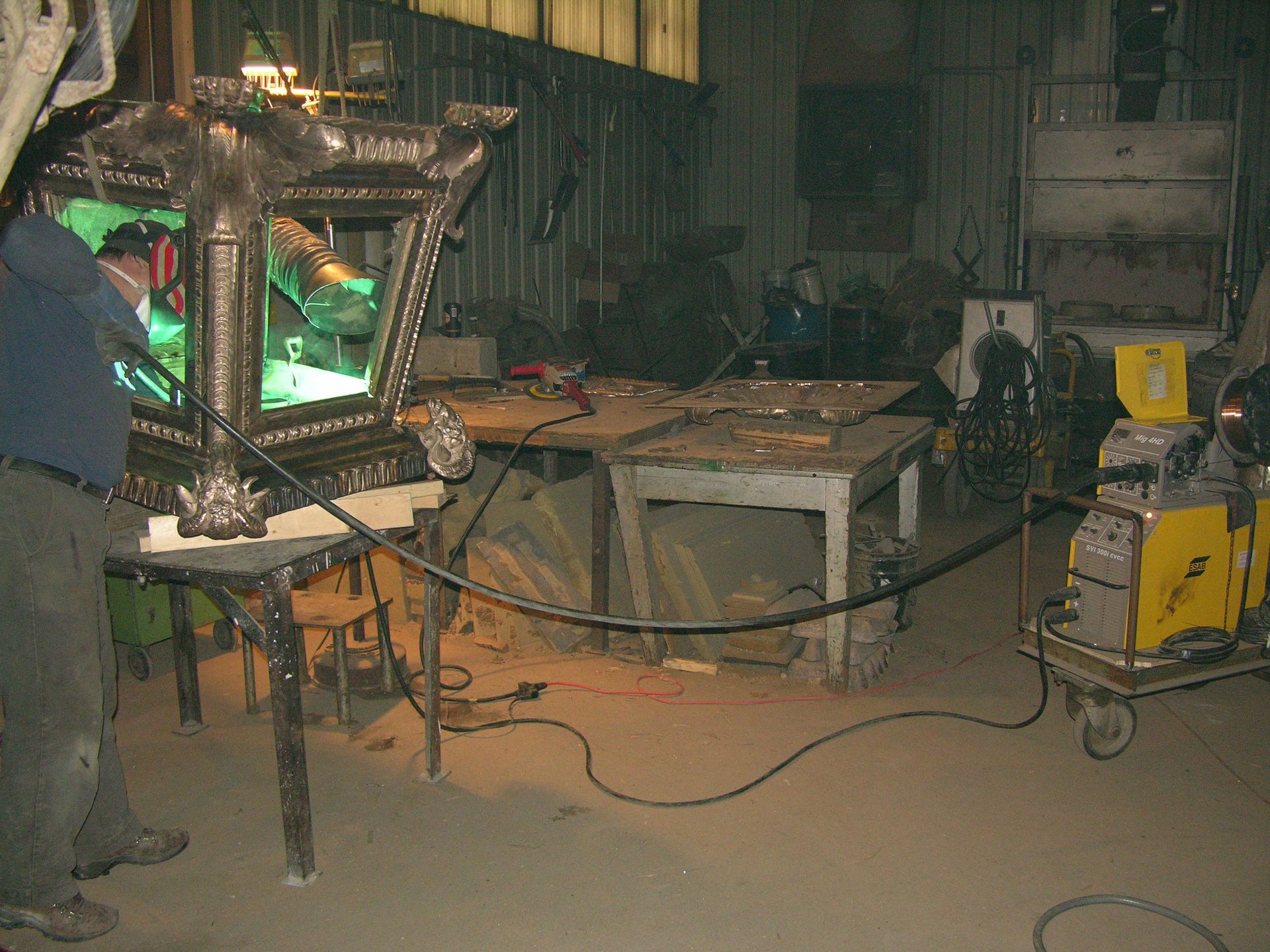 Cluttered workshop with a framed mirror on a table, cords on the floor, and yellow equipment on the right.