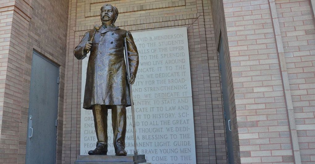 A bronze statue of a person in a coat stands before a brick wall with an inscribed stone plaque.