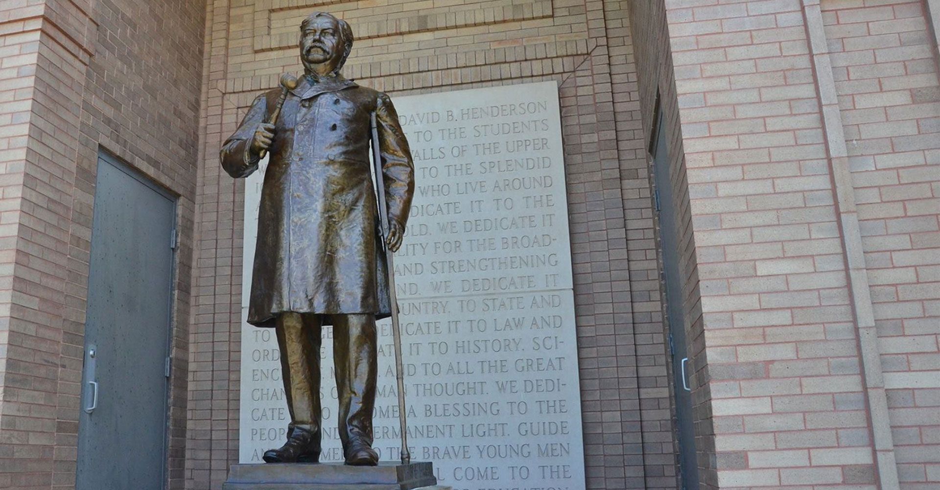 A bronze statue of a person in a coat stands before a brick wall with an inscribed stone plaque.
