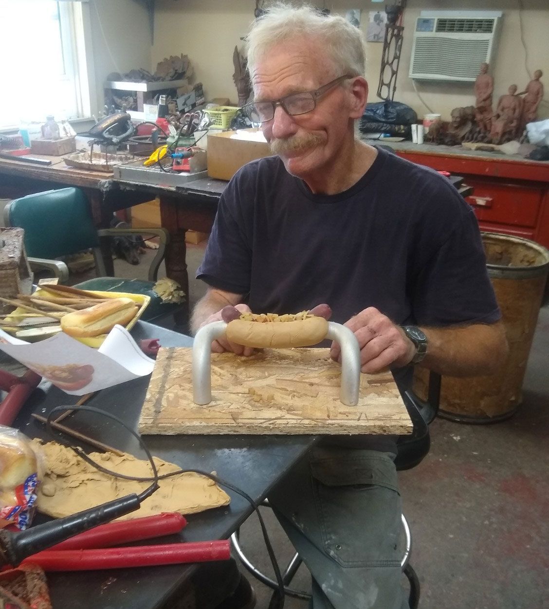 An individual uses a tool to apply a thick, tan material onto a curved metal handle on a wooden board in a workshop.
