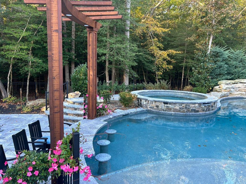 Backyard pool with stone spa, wooden pergola, chairs, pink flowers, and wooded garden backdrop