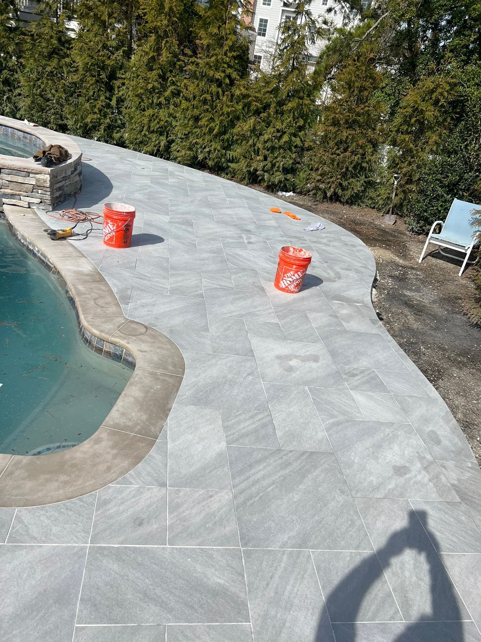 Patio with blue-gray pavers, curved pool, orange buckets, and evergreen trees in the background.