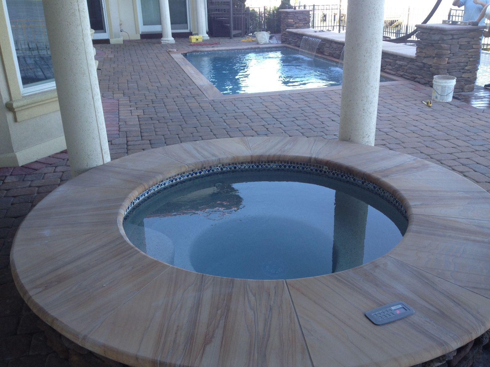 A circular hot tub with a stone surround, patio and swimming pool. Outdoors with pillars.