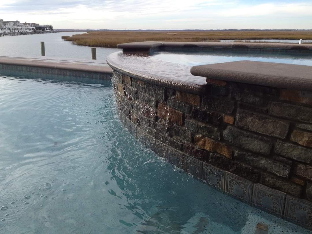 Stone-walled pool with water cascading into a larger body of water, cloudy sky overhead.