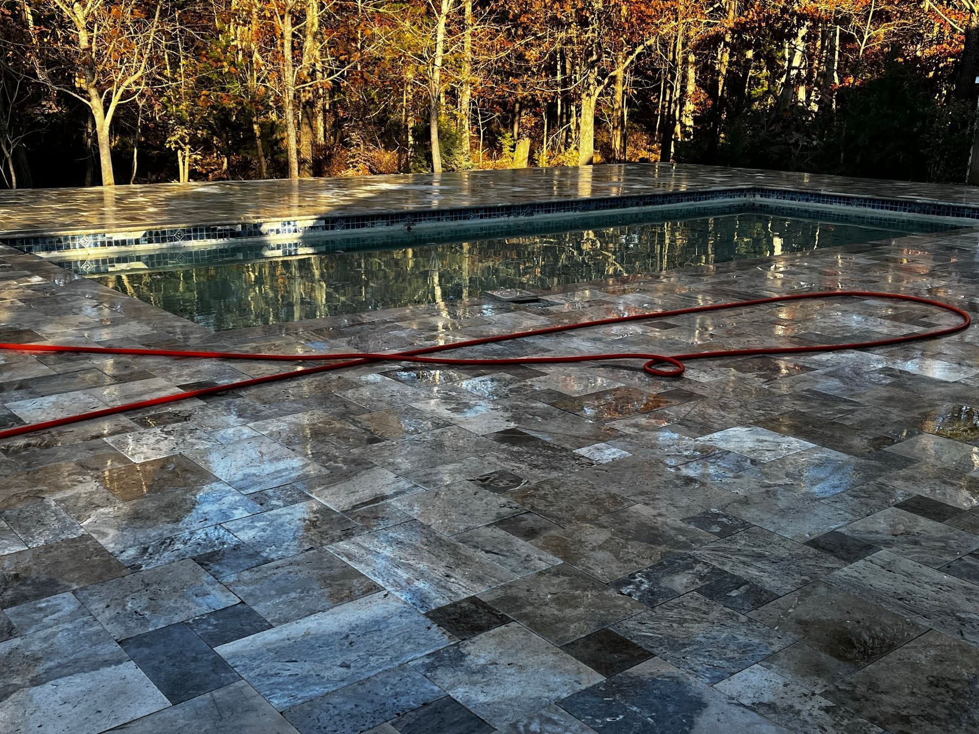 Wet stone patio with a red hose by a rectangular pool, surrounded by trees.