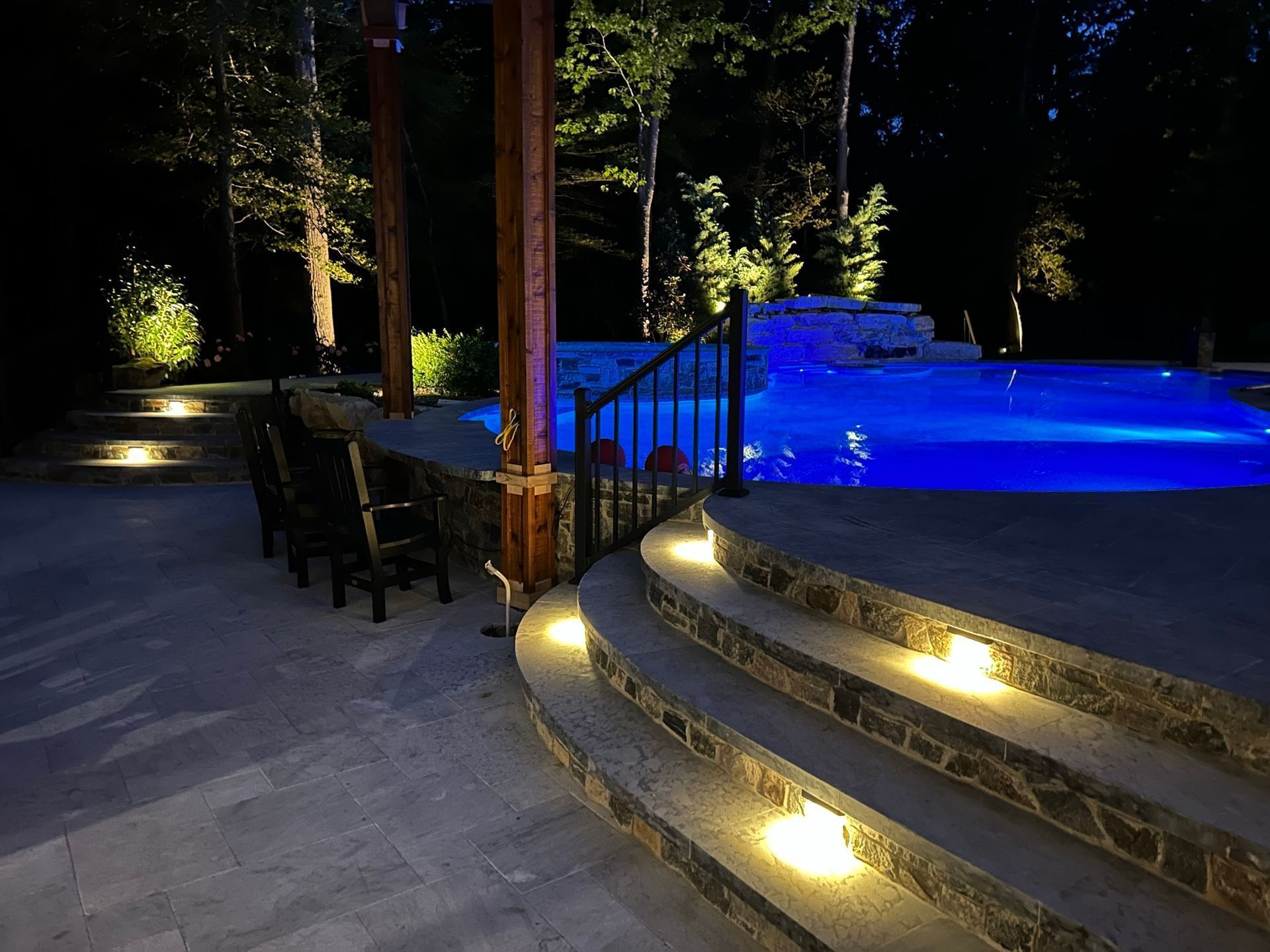 Outdoor patio with illuminated steps leading to a blue-lit pool at night. Trees and wooden pergola frame the scene.