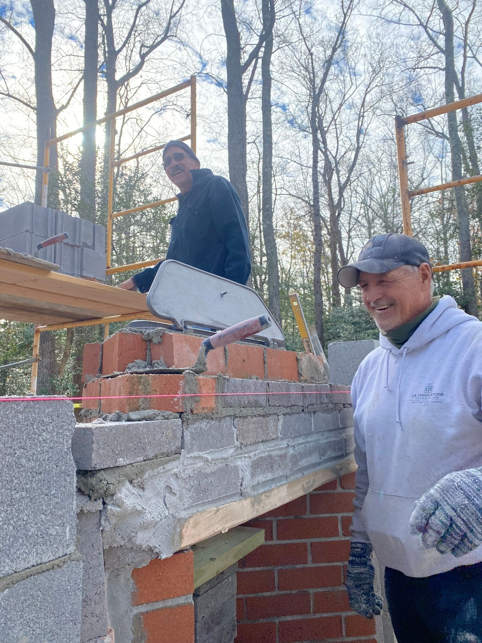 Two construction workers smiling while building a brick structure outdoors.