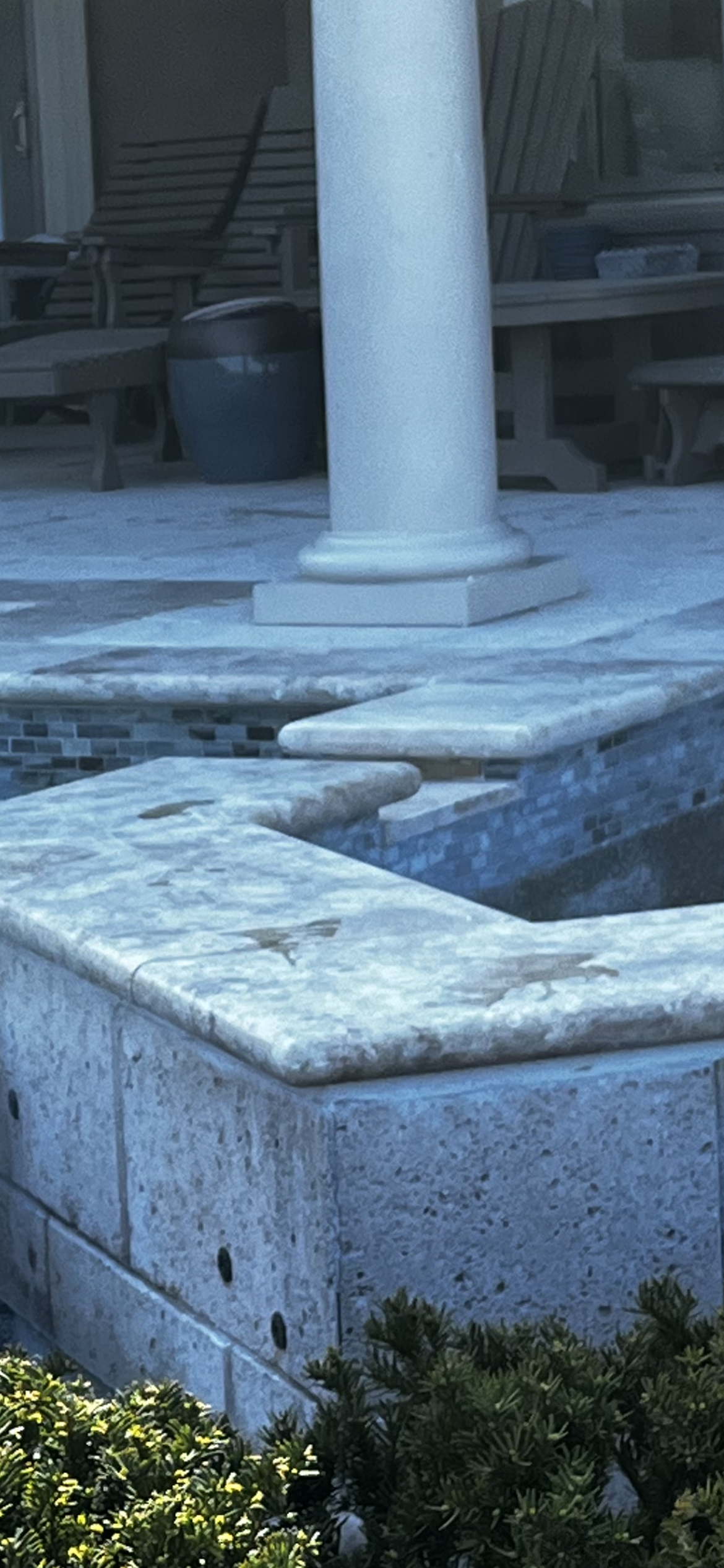 A marble structure with a column and a water feature. Greenery in the foreground.