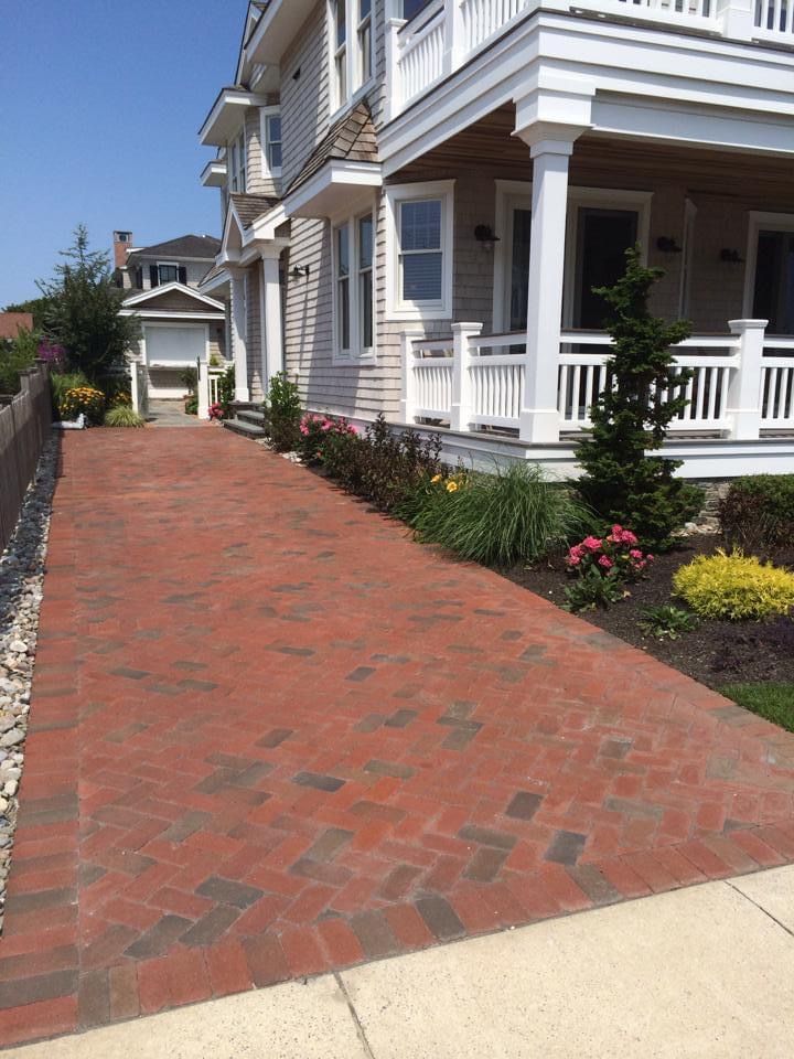 Brick driveway leading to a light-colored house with a porch and landscaping. Sunny day.
