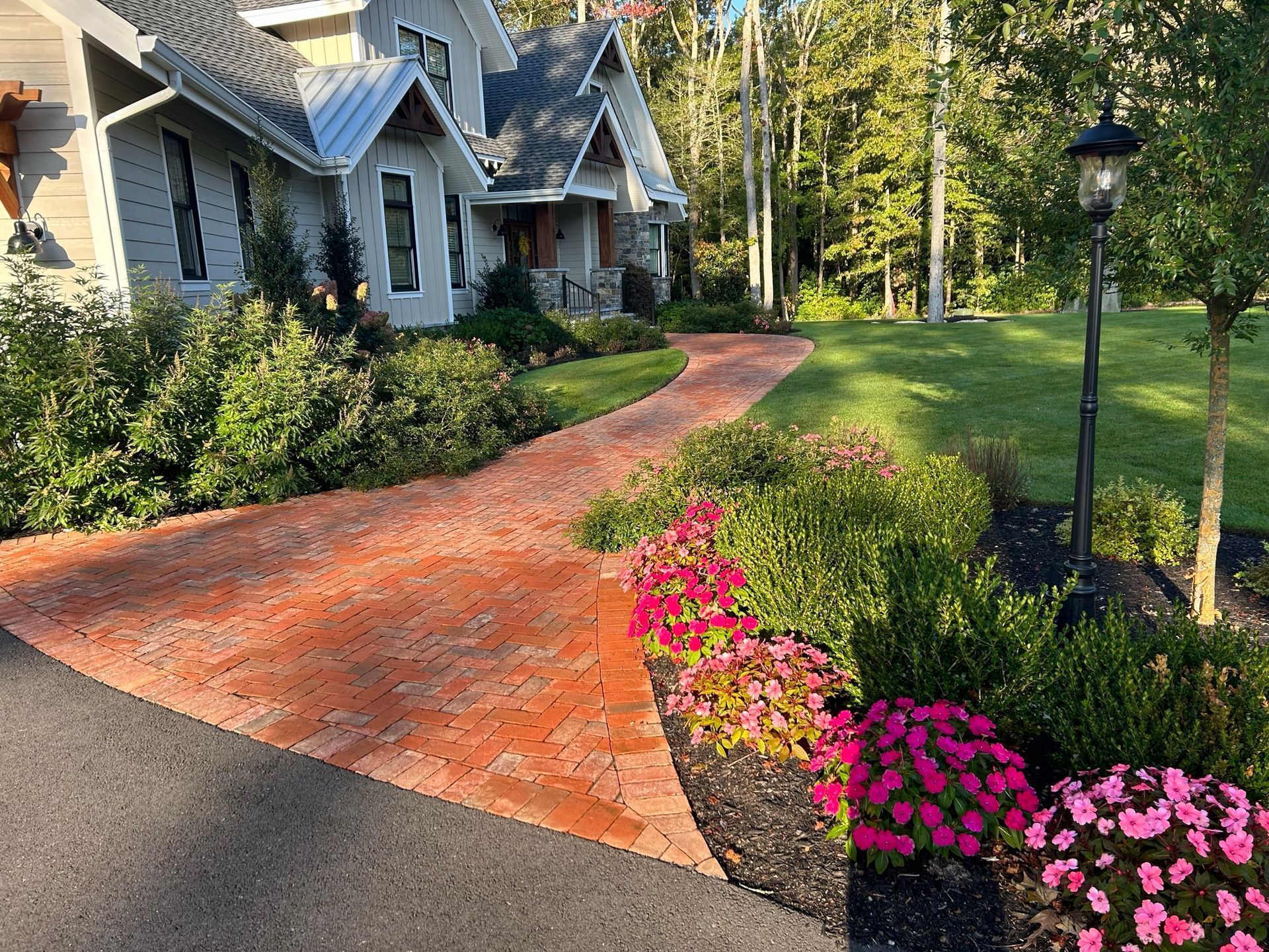 Brick pathway leads to a house with a green lawn and flower beds, bordered by a black driveway.