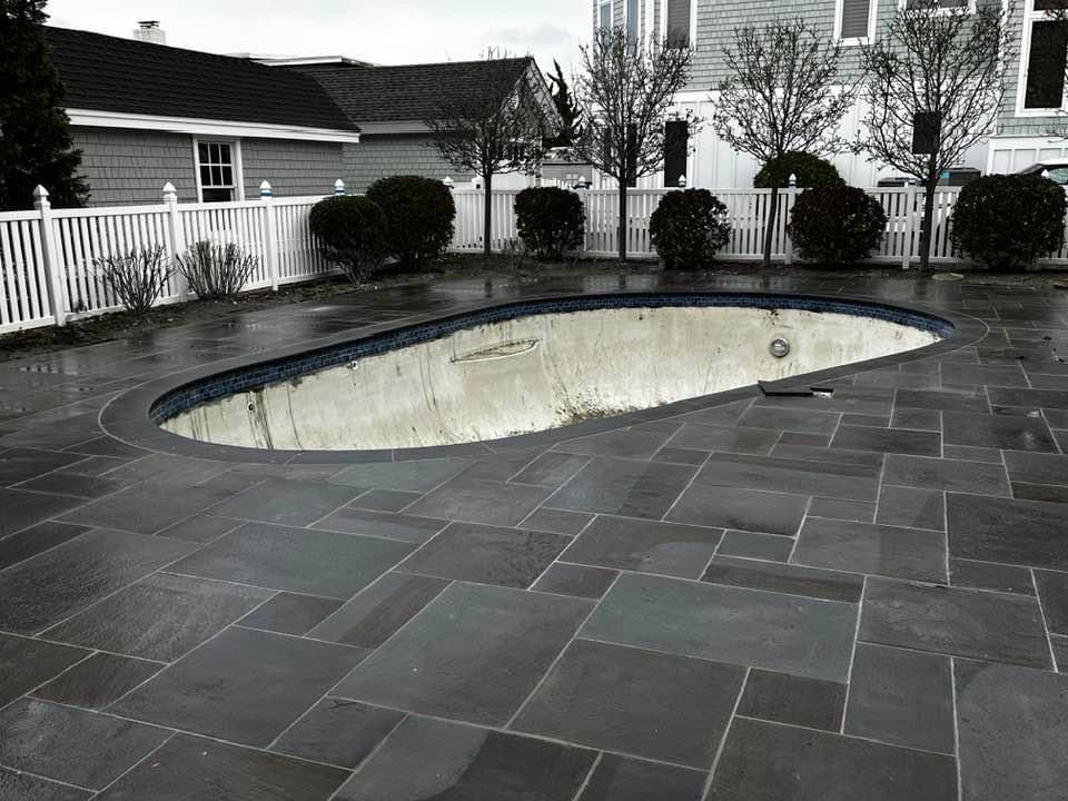 Empty pool surrounded by dark paving stones, a white fence, and houses on a cloudy day.