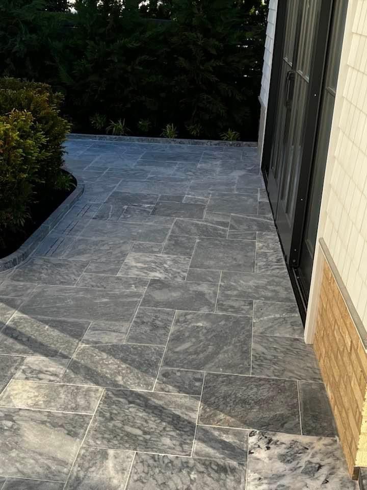 Stone patio with dark grey tiles next to a building and greenery.