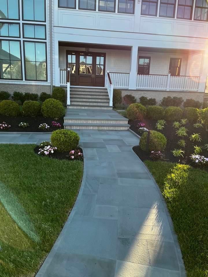 Stone walkway leads to house with steps, surrounded by green grass, bushes, and flowerbeds.