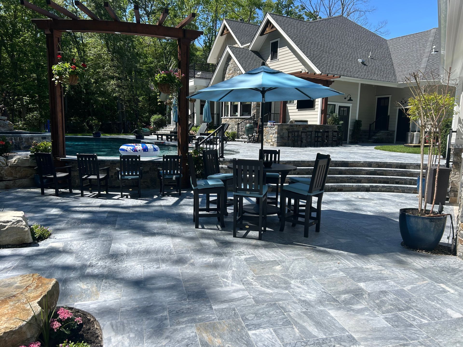 Patio with outdoor furniture, pool, pergola, and house with blue umbrella on a sunny day.