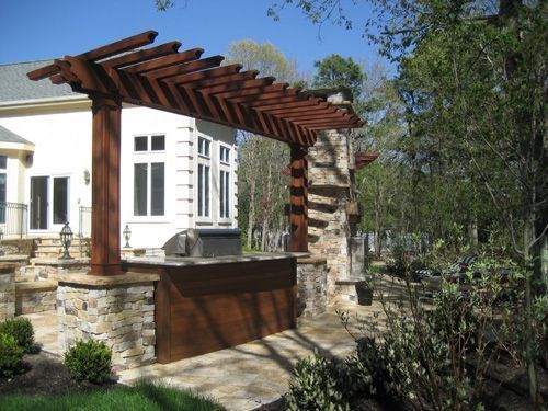Wooden pergola over outdoor kitchen with stone base, next to a house and garden.