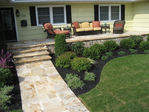 Stone walkway leads to a yellow house with a porch. Landscaping includes shrubs and lawn. Outdoor furniture on the porch.