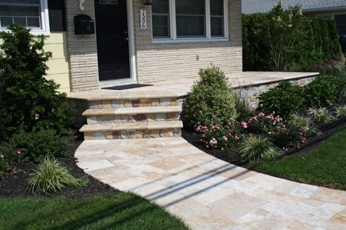 Stone walkway leading to a house with steps and landscaped garden, featuring beige stone and green plants.