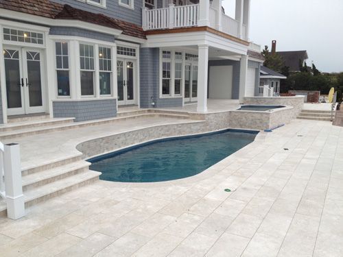 A luxury home with a swimming pool and patio. Light stone flooring, blue pool water, and white railings.