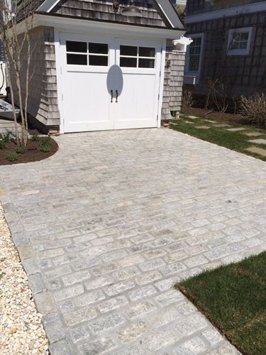Gray brick driveway leading to a white garage door, surrounded by grass and landscaping.