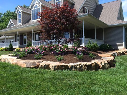 House with wrap-around porch and landscaped flowerbed edged with large rocks and a red tree.