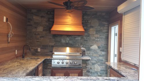 Outdoor kitchen with granite countertops, stone backsplash, and built-in grill under a wooden hood.