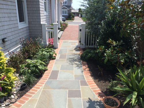 Brick-lined walkway with stone tiles leading to a white gate, flanked by greenery and a beach house.