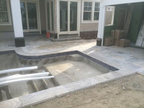 Unfinished pool area with gray stone patio, steps, and blue tile trim. Building and boxes in background.