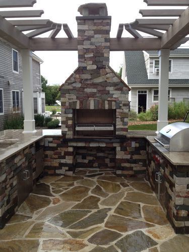 Outdoor kitchen with stone accents, a built-in grill, and fireplace, under a wooden pergola on a flagstone patio.