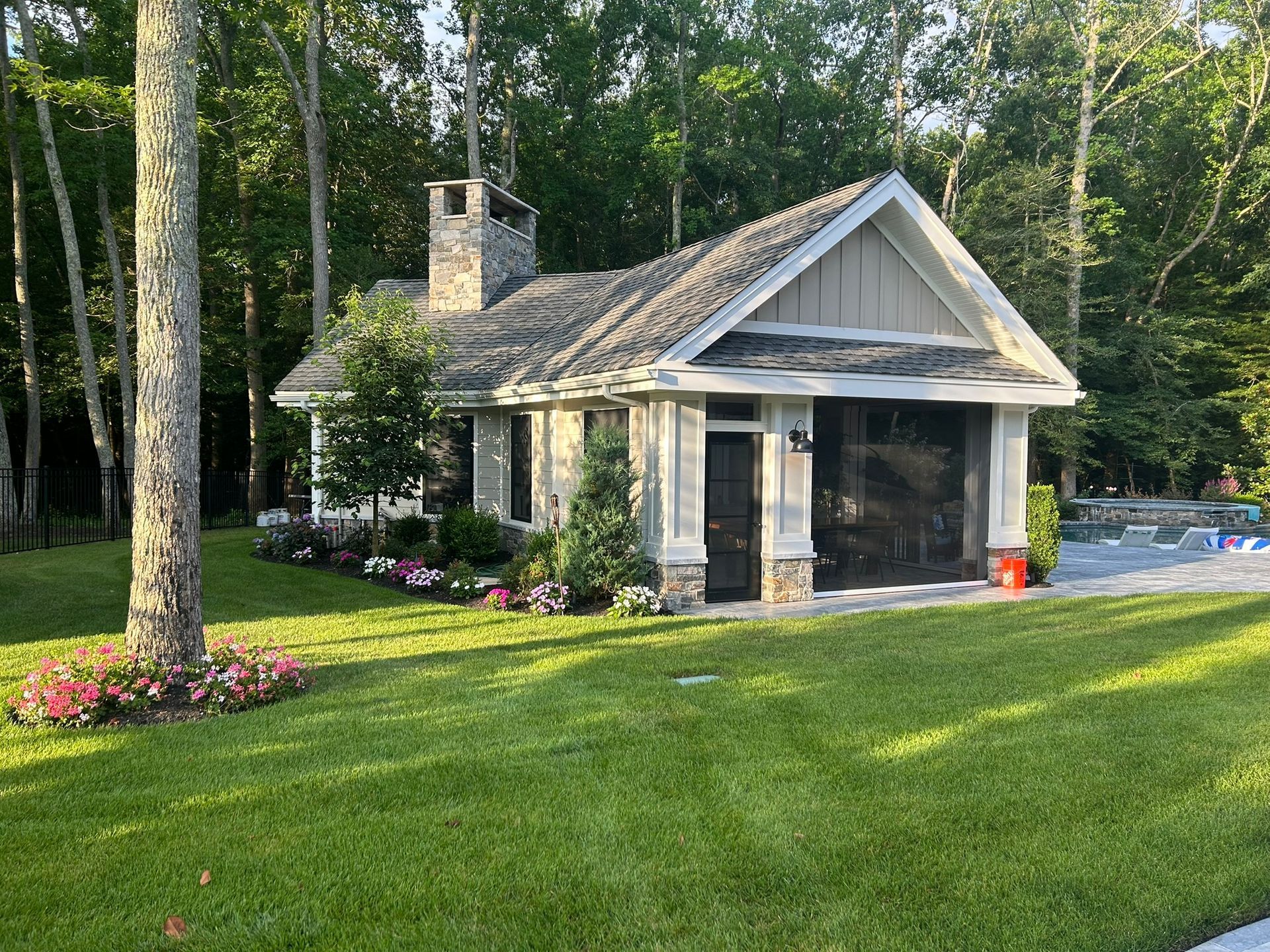 Small cottage with stone chimney and screened garage in a lush green yard, surrounded by trees.