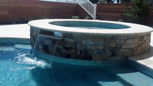 Stone-faced hot tub with water cascading into a pool. White coping, wooden wall, steps.