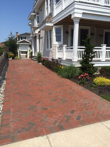 Brick driveway leading to a light-colored house with a porch and landscaping.