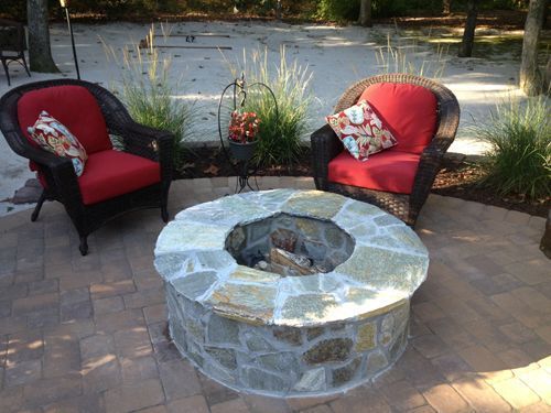 Fire pit with stone surround, two wicker chairs with red cushions, outdoor patio setting.