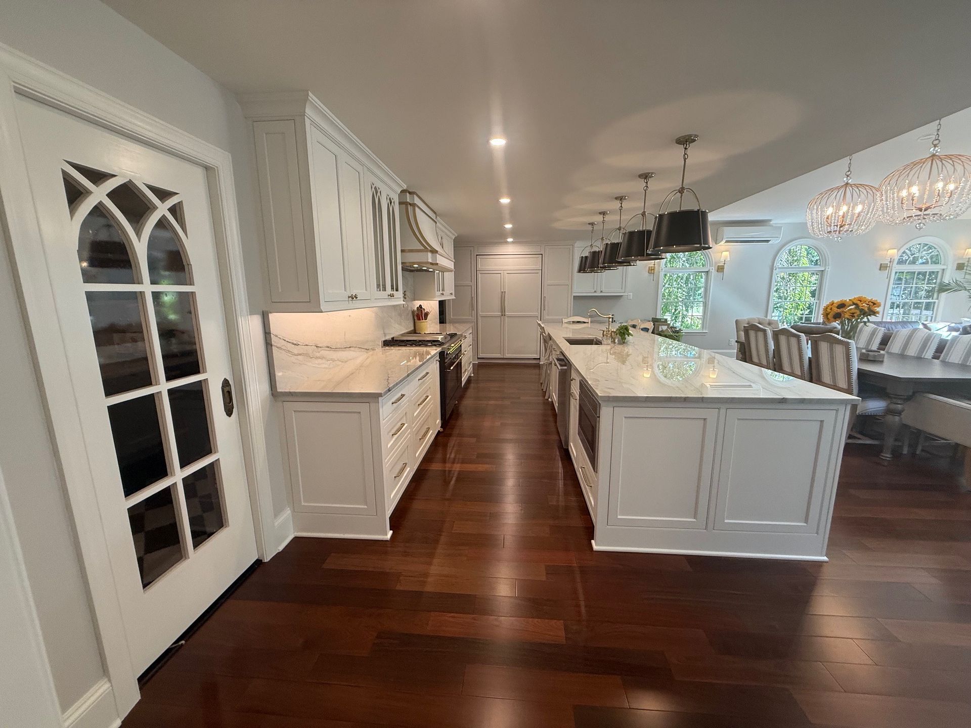 White kitchen with dark wood floors, island, cabinets, and arched-glass door. Dining area visible.