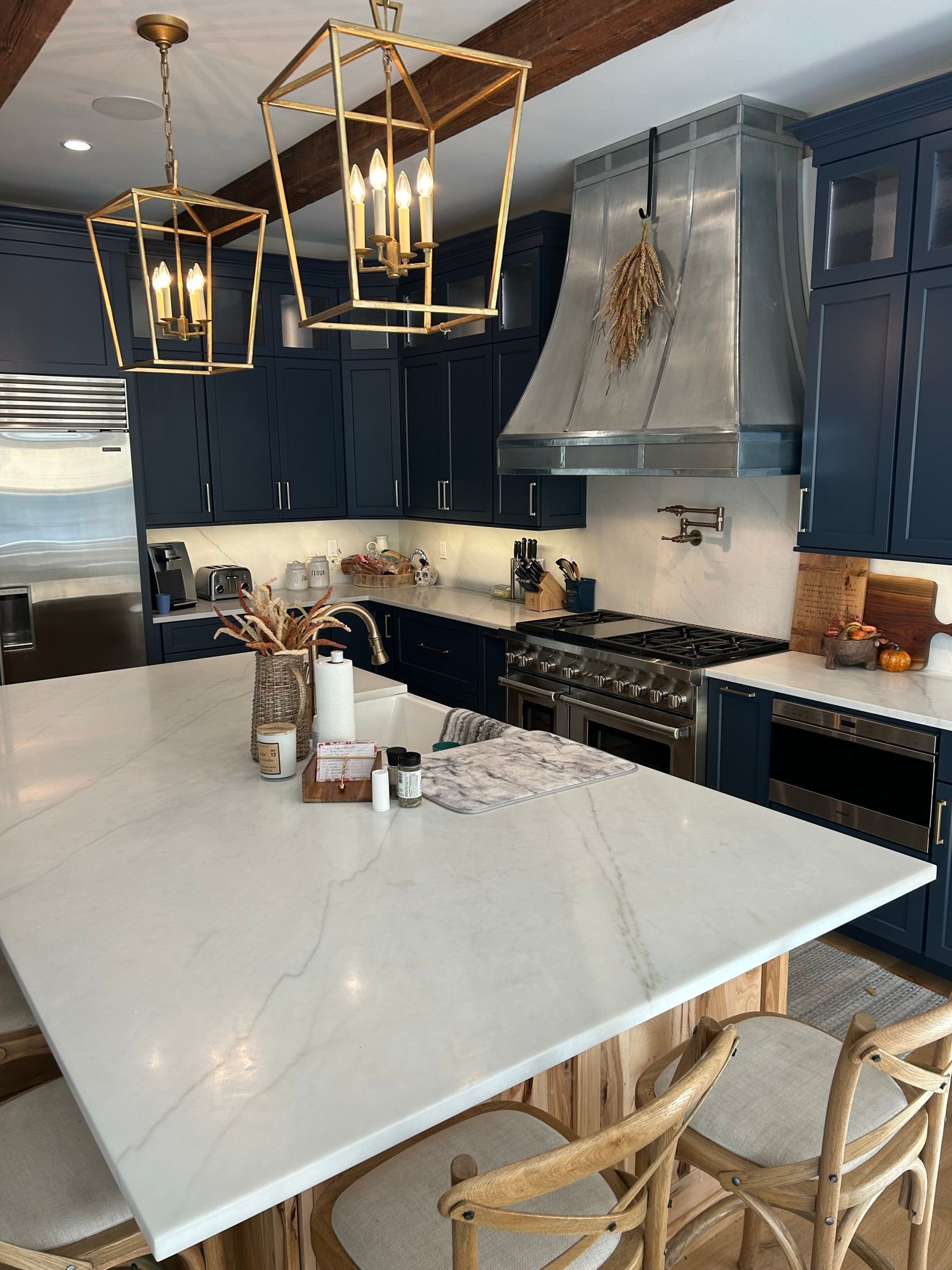 Blue kitchen with white countertop island, gold light fixtures, stainless steel range hood, and wooden beams.