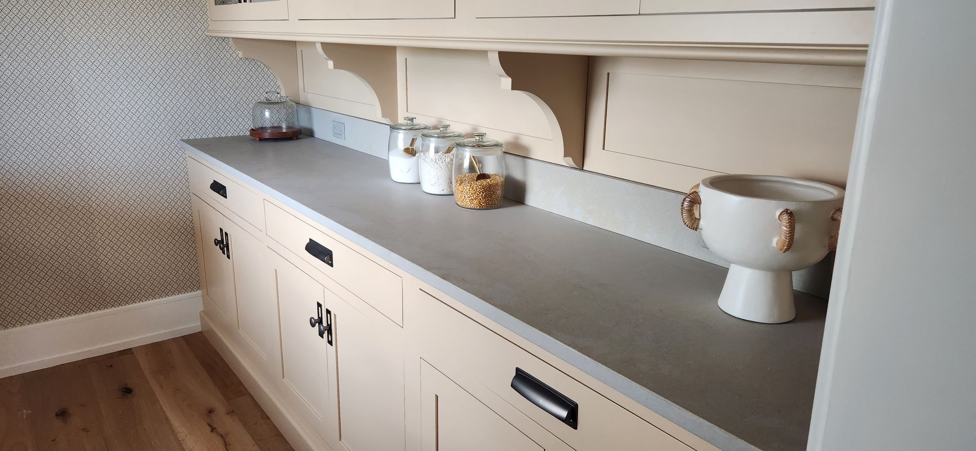 Kitchen counter with cabinets, jars, and a decorative bowl. Gray countertop with white cabinets and a patterned wallpaper background.