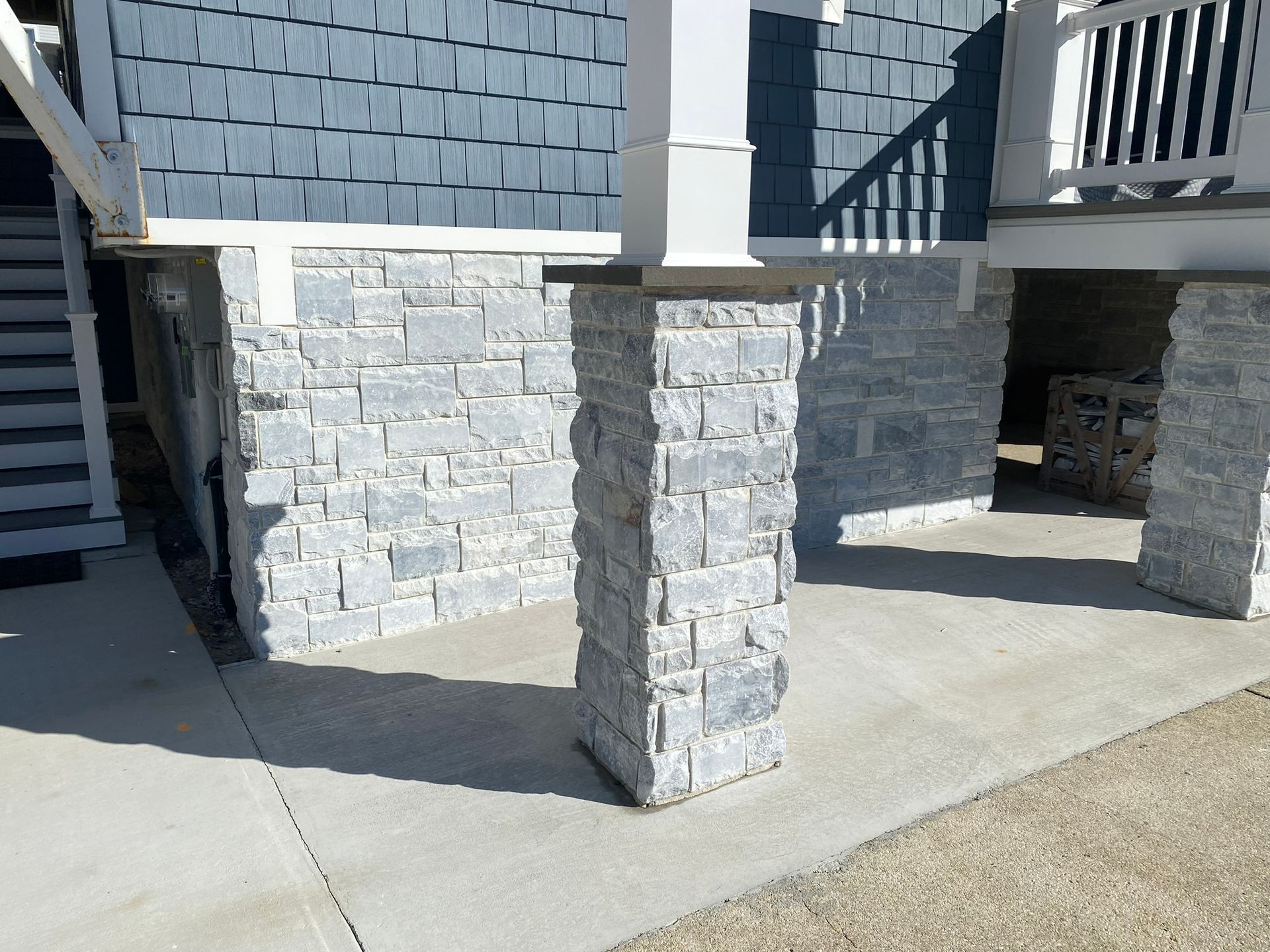 Stone-faced supports holding up a blue shingled house. Concrete slab and gravel ground. Bright daylight.
