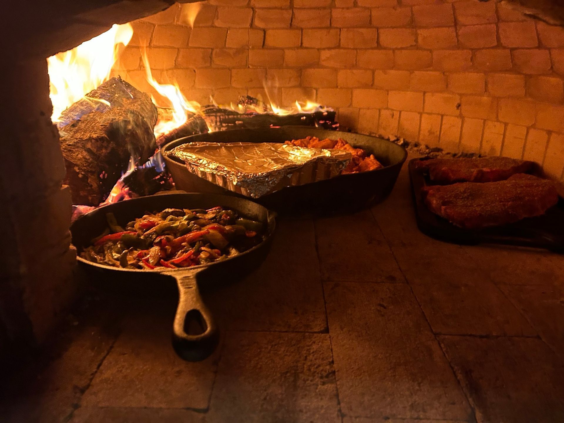 Food cooking in a brick oven with a fire. Includes cast iron skillets and other dishes.