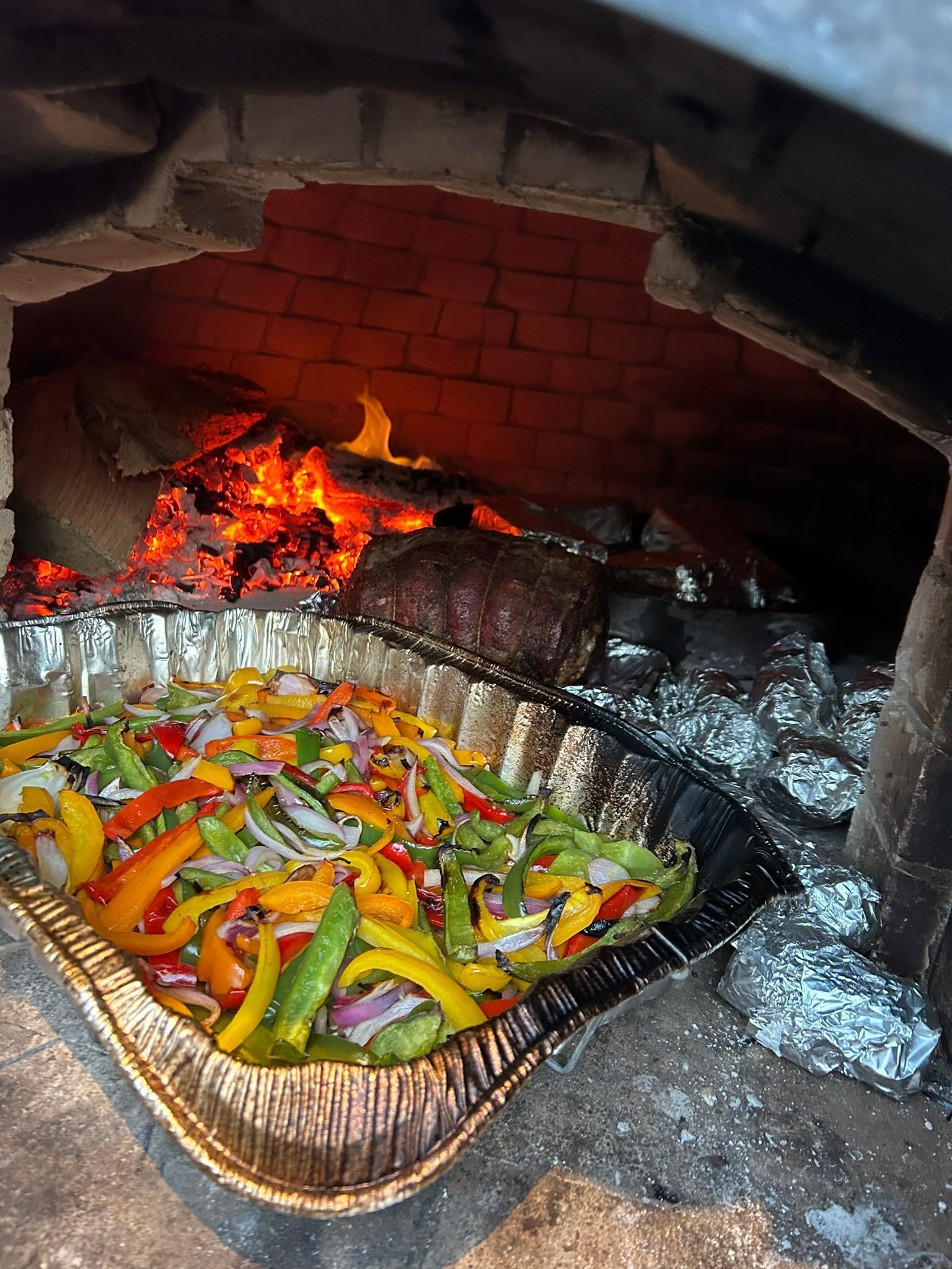 A metal pan of sliced peppers and onions cooking in a wood-fired brick oven with a glowing fire.