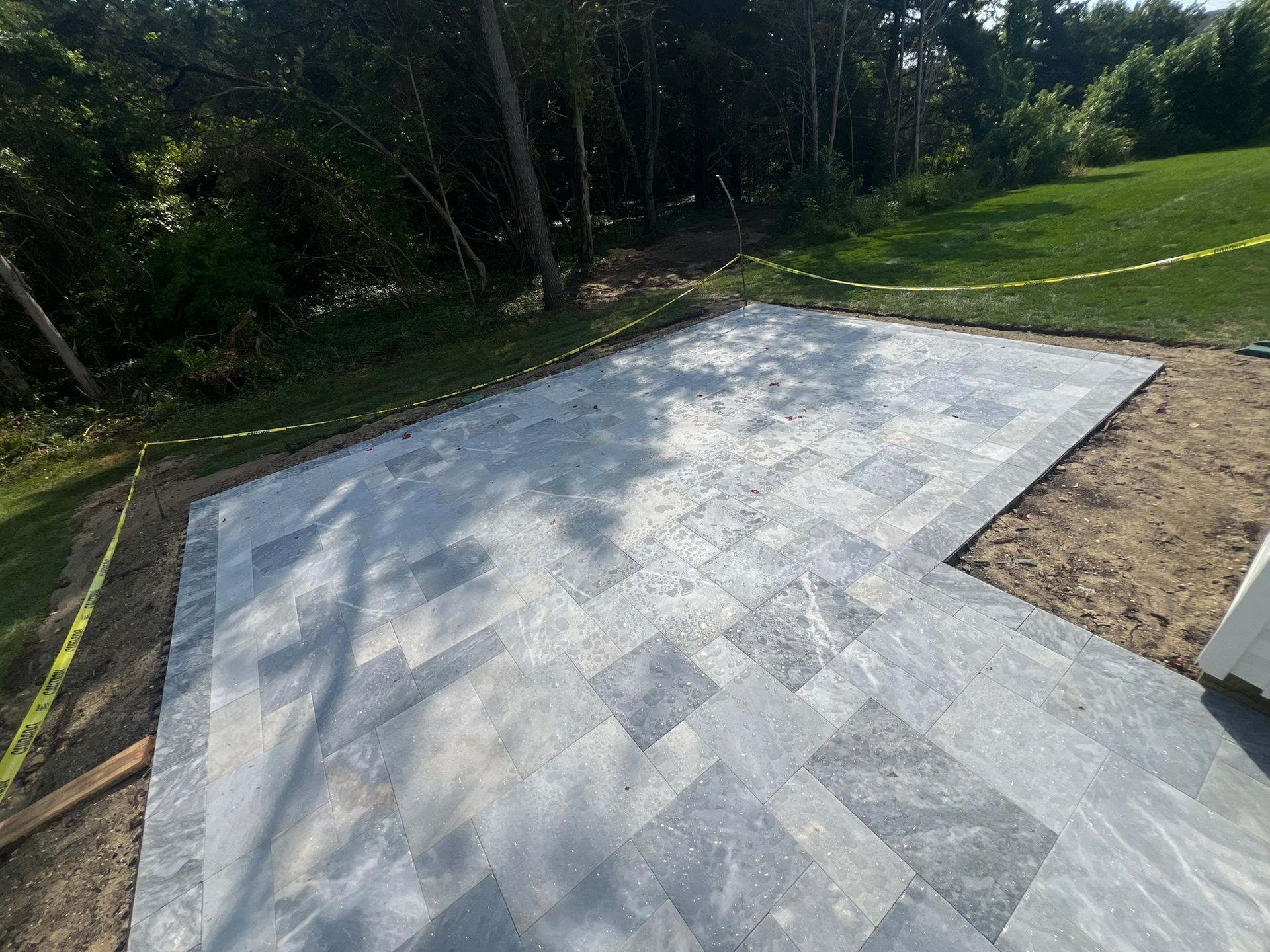 Gray stone patio under construction, bordered by grass, trees, and safety tape.