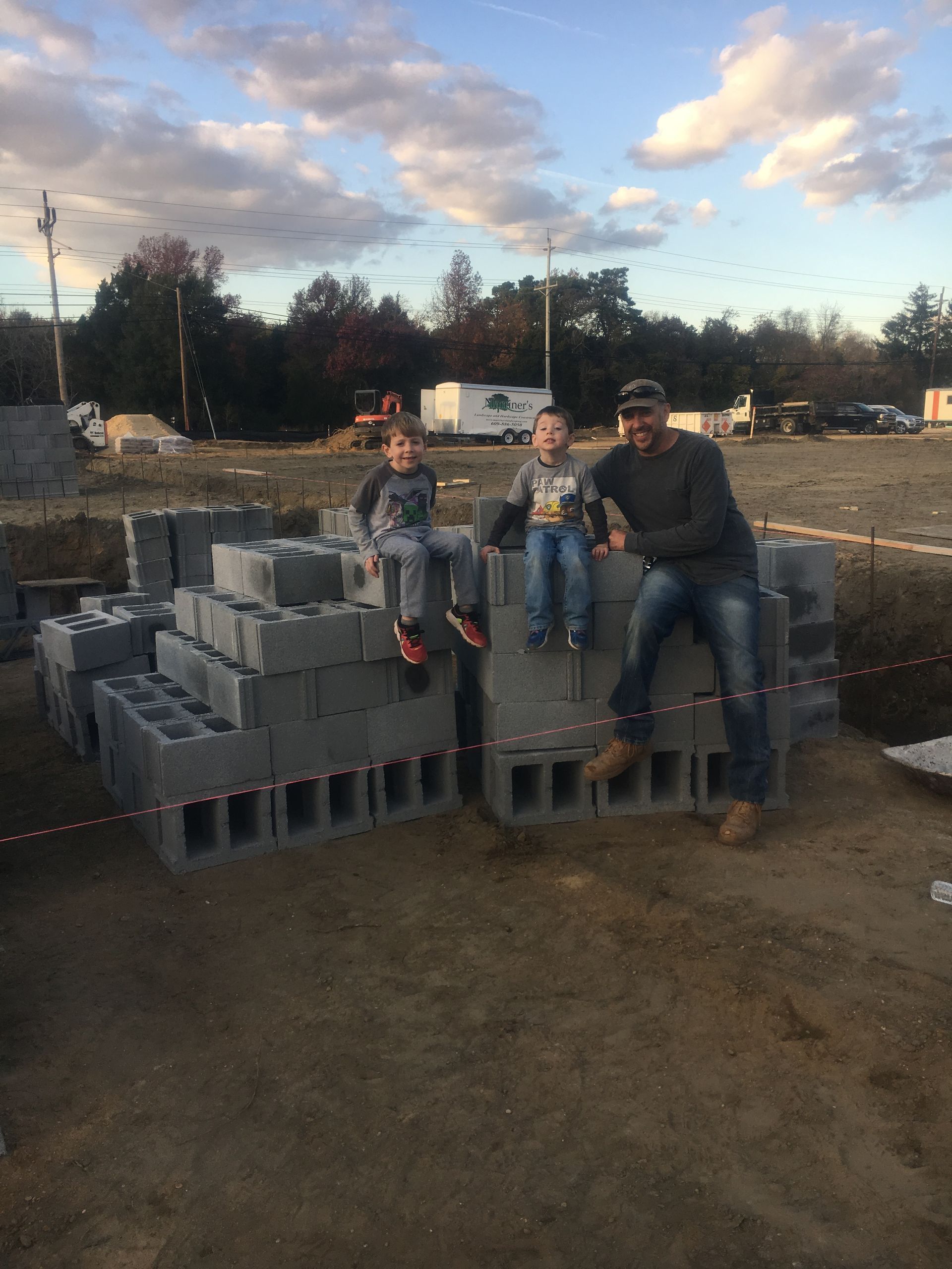 A man and two children sit on concrete blocks at a construction site; overcast sky.