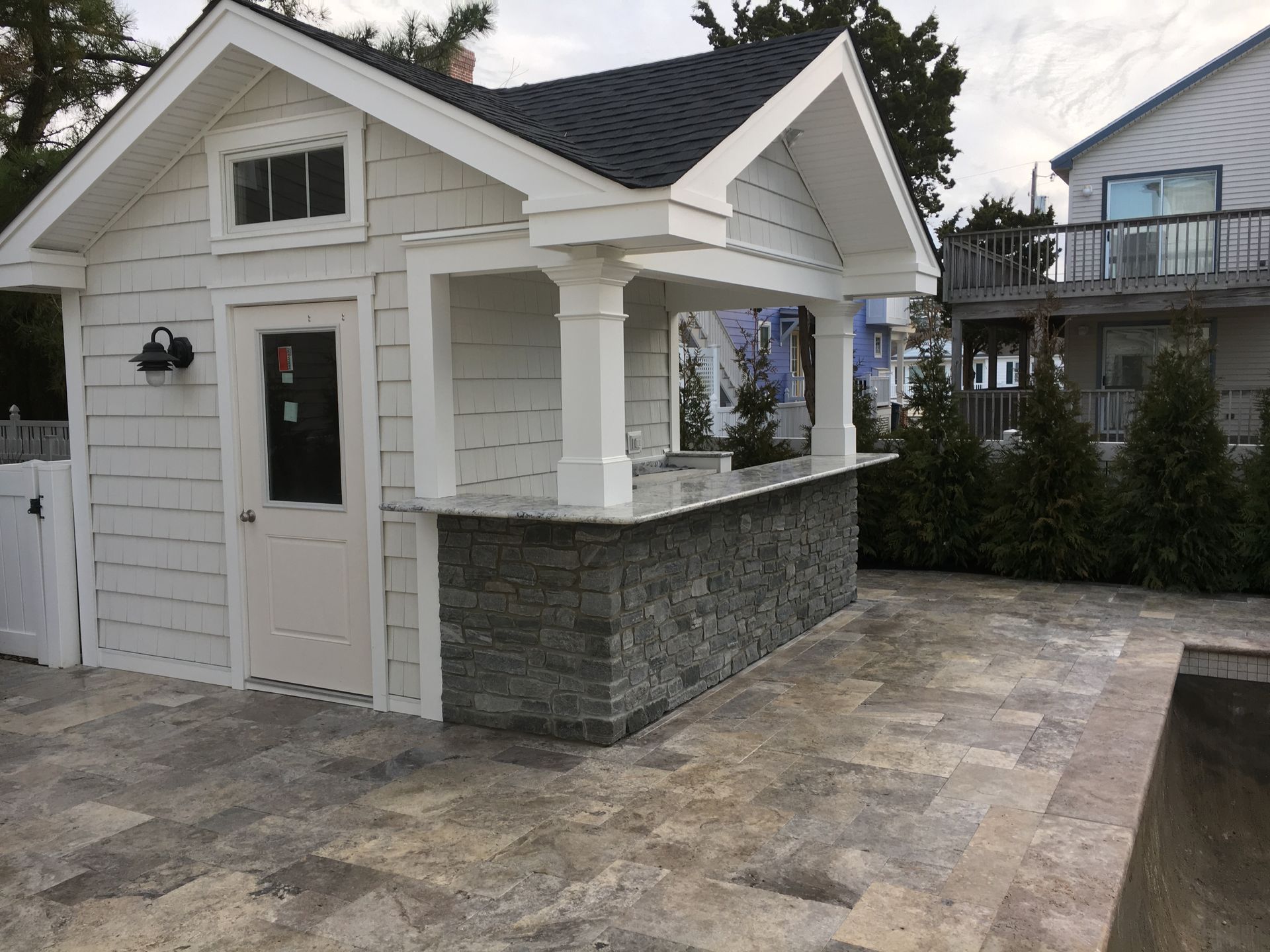 White outdoor bar with stone base, gray countertop, and attached shed with a black roof.